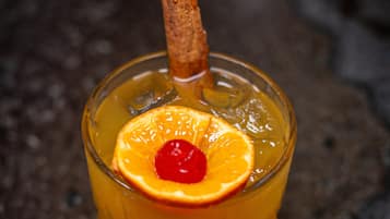 A glass of amber-colored beverage with a slice of orange and a cherry, garnished with a cinnamon stick, on a textured surface.