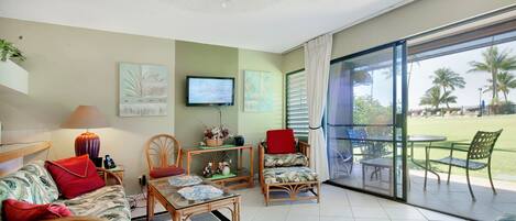 A spacious living room with a sofa, chairs, a coffee table, and a TV. There is a balcony with outdoor furniture and palm trees visible through the glass doors.