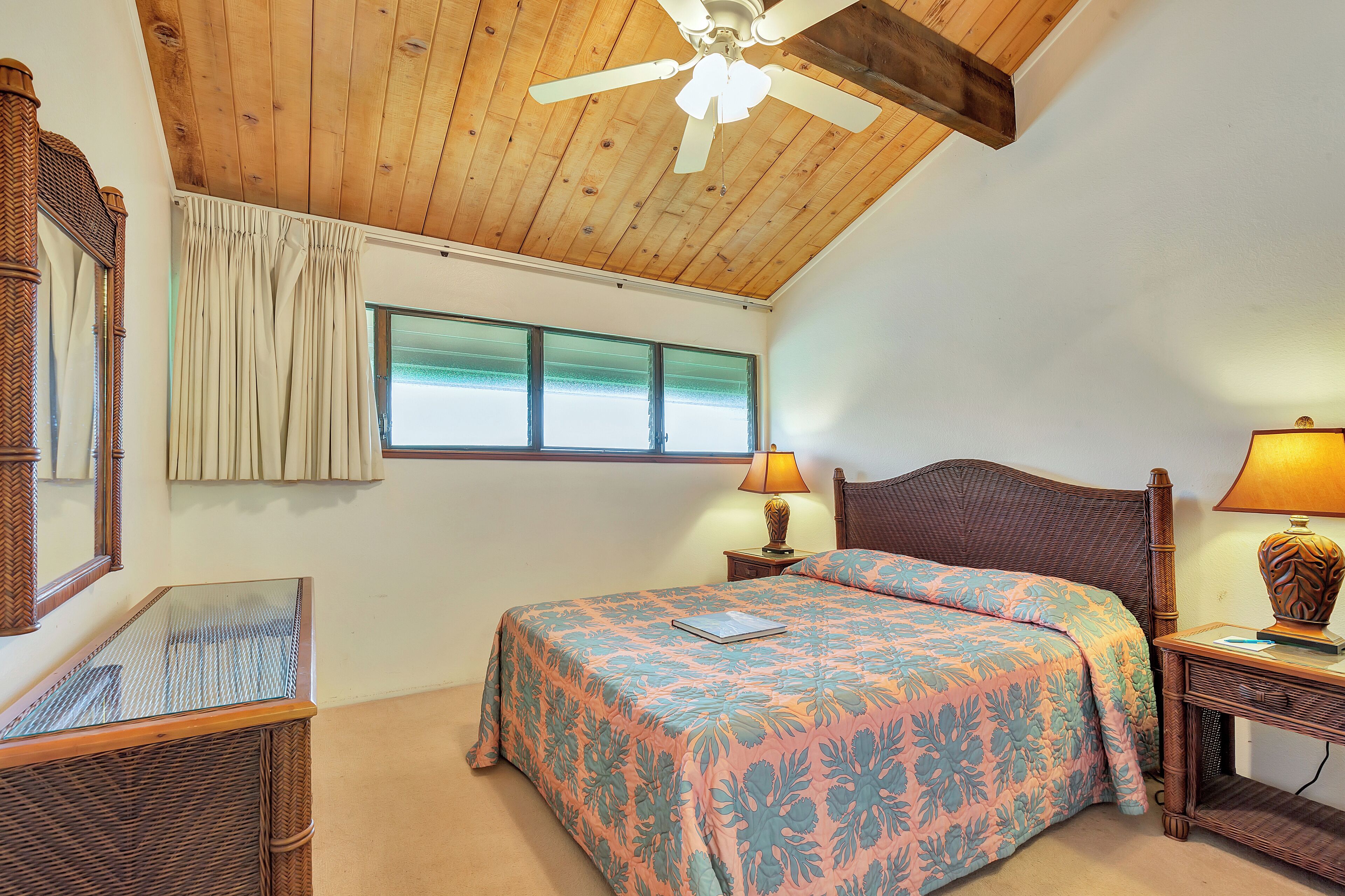 Un dormitorio con techo de madera, una cama con colcha estampada, una mesita de noche con lámpara y una ventana con cortinas.