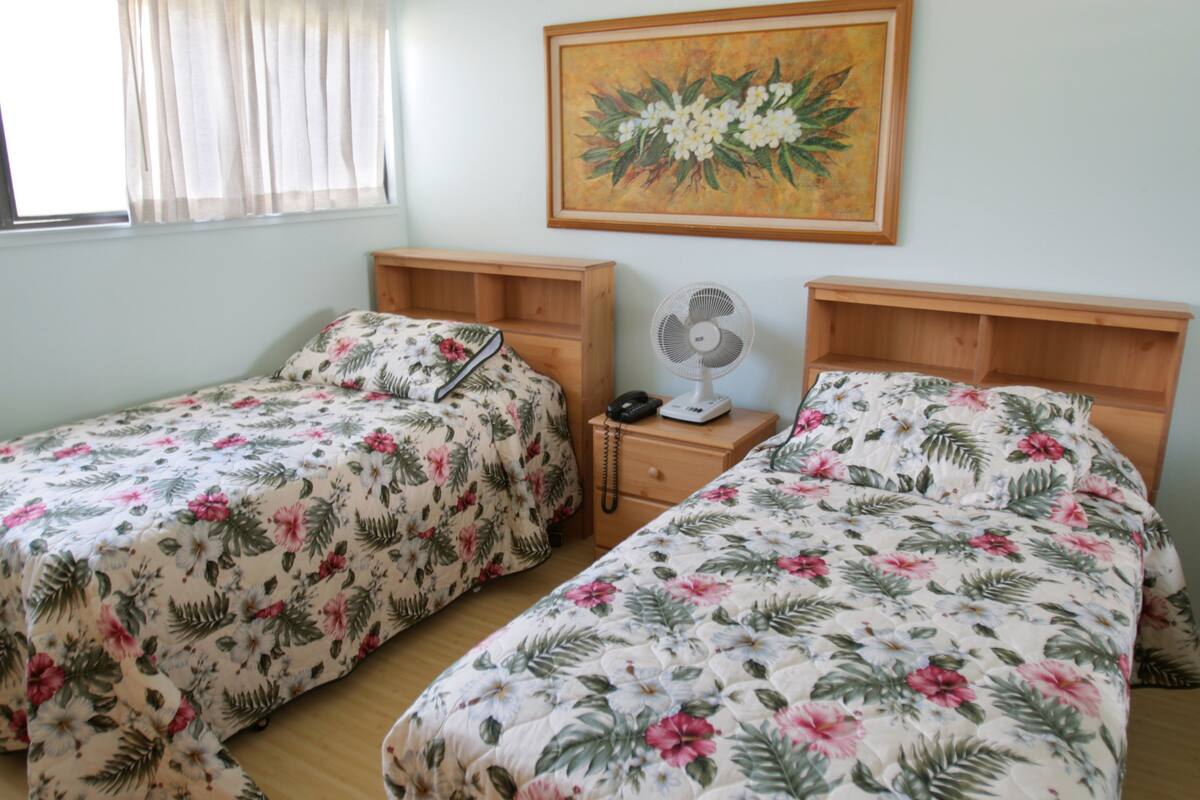 Habitació amb dos llits individuals, cadascun amb una colcha floral i amb els reposacapses de fusta. Hi ha una tauleta de nit amb un ventilador i un telèfon entre els llits. Un quadre penja a la paret sobre els llits.
