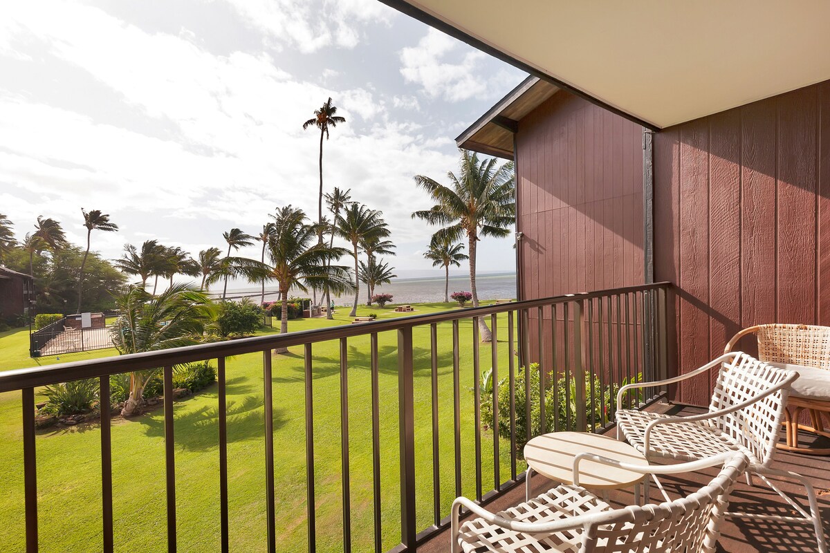 A balcony with a view of a lawn and palm trees, a white wicker chair, and a table.