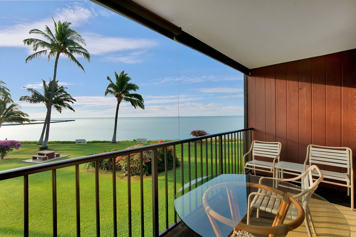 A balcony with a glass table and chairs, overlooking a well-maintained lawn with palm trees and a view of the ocean.