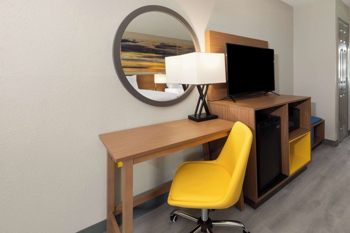 A hotel room with a wooden desk, a yellow chair, a flat-screen TV, a lamp, and a microwave.