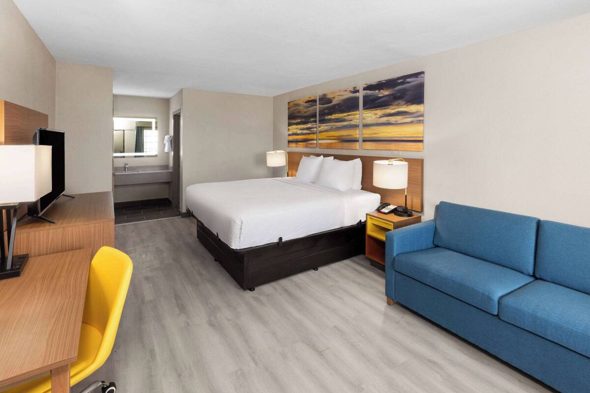 A hotel room with a bed, a desk with a chair, a blue sofa, and a painting on the wall.