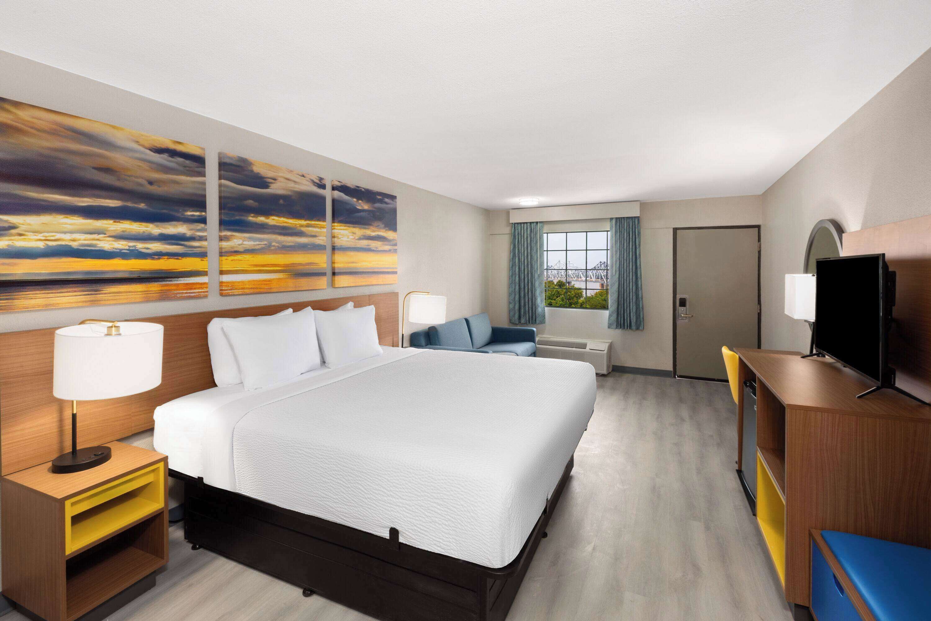 Una habitación de hotel con una cama grande, un escritorio, un televisor y un cuadro de una puesta de sol.