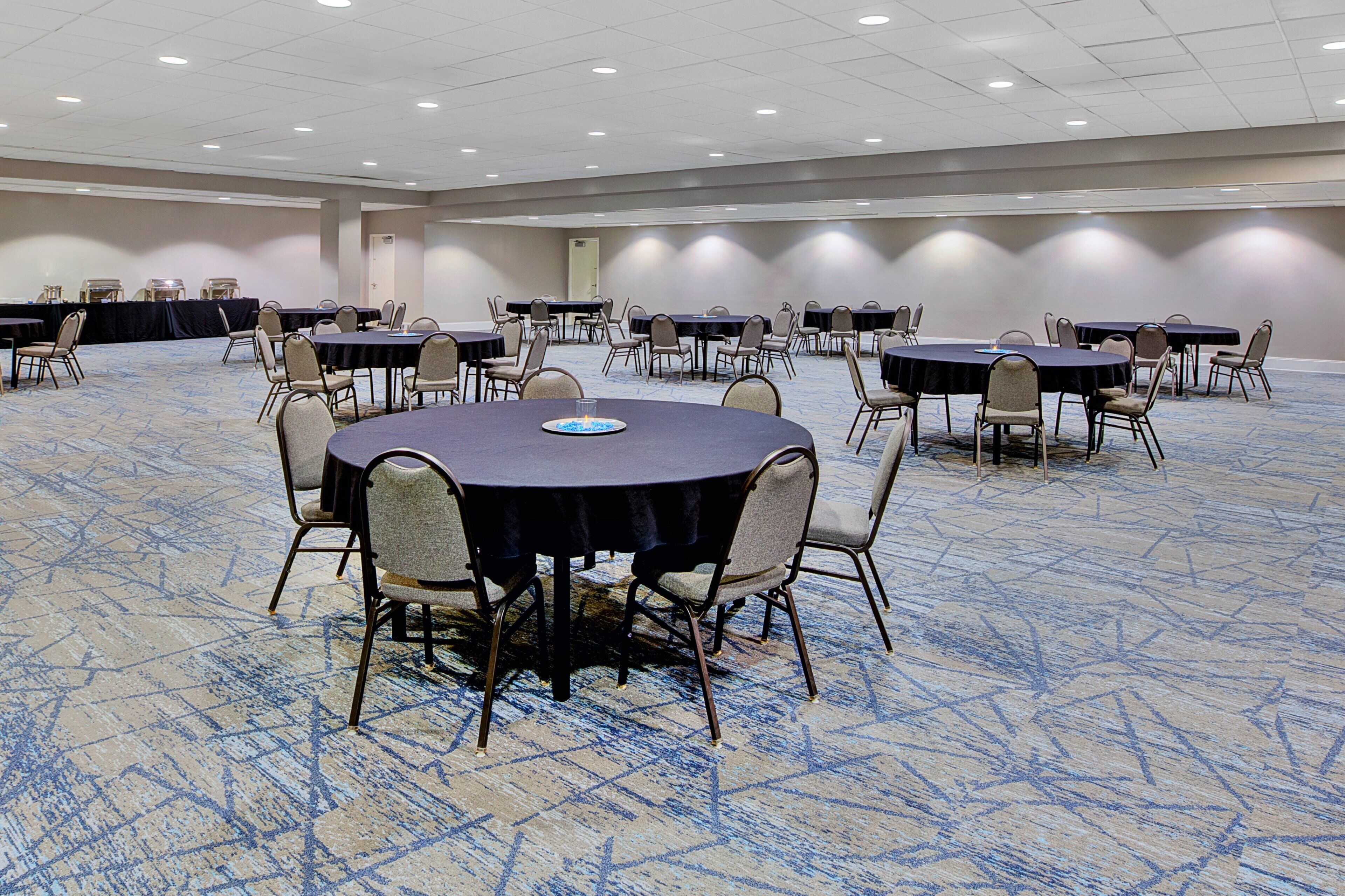 Un salón de conferencias con mesas y sillas redondas, piso alfombrado y una puerta al fondo.