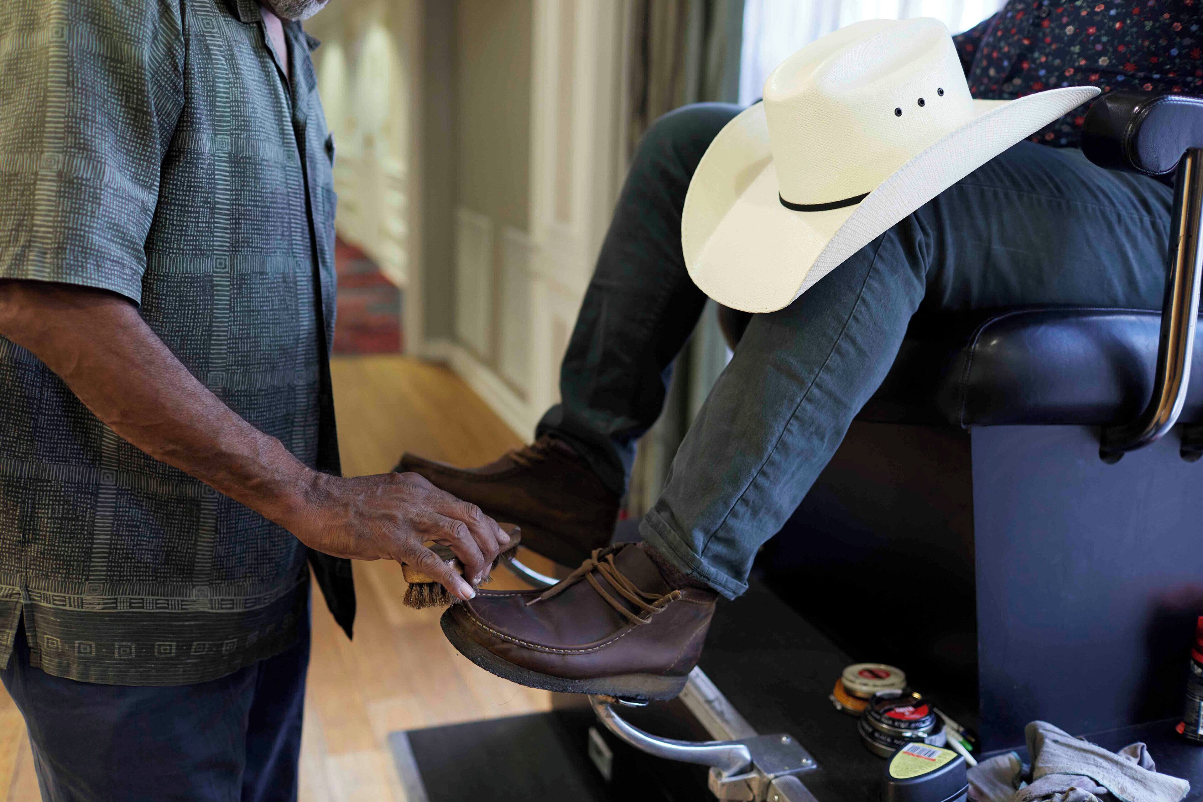 Una persona sentada en un sofá de cuero negro, usando un sombrero vaquero blanco con una carita sonriente, y otra persona de pie a su lado, sosteniendo un zapato de cuero marrón.