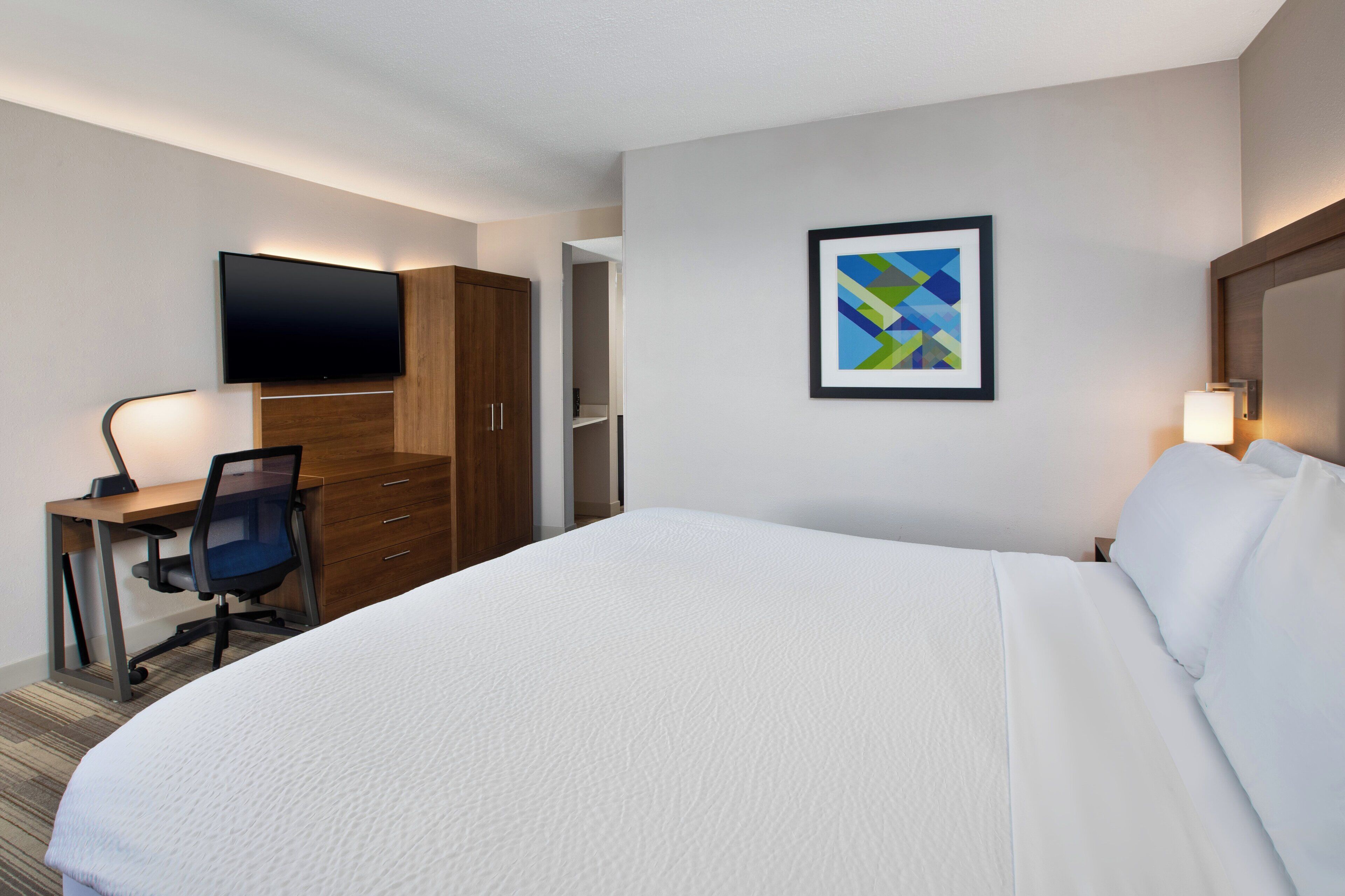 A hotel room with a large bed, a desk with a chair, a flat-screen TV mounted on the wall, and a framed abstract painting.
