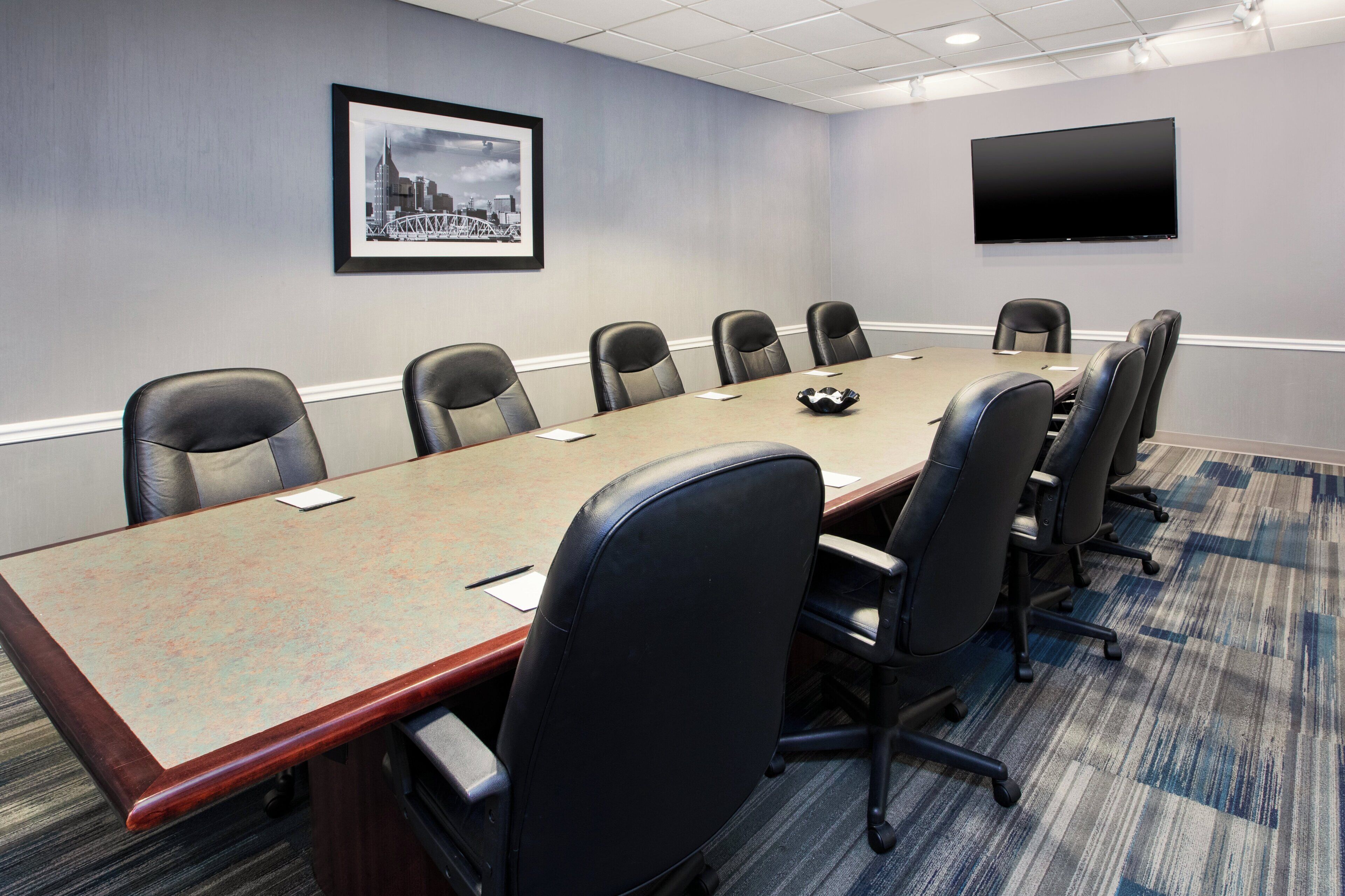 A conference room with a long table, six chairs, a mounted TV, and a framed cityscape picture on the wall.