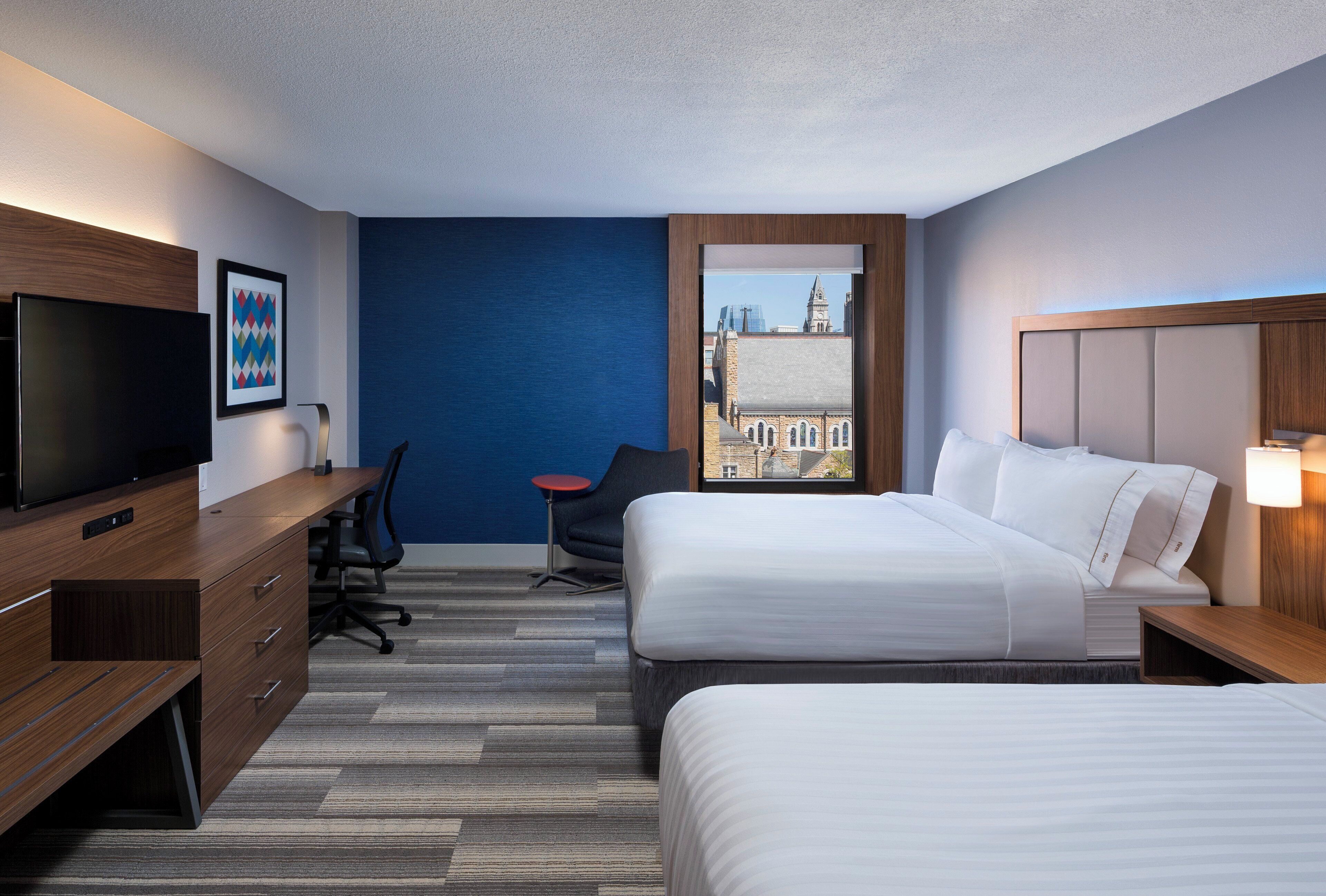 A hotel room with two beds, a desk, a chair, a television, and a window with a city view.