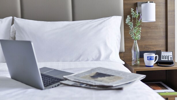Un lit bien fait, avec du linge de lit blanc, un ordinateur portable posé sur le lit, un journal, un vase rempli de fleurs et une table de chevet avec une lampe et un téléphone.