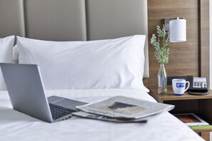 A neatly made bed with white linens, a laptop on the bed, a newspaper, a vase with flowers, and a bedside table with a lamp and a phone.