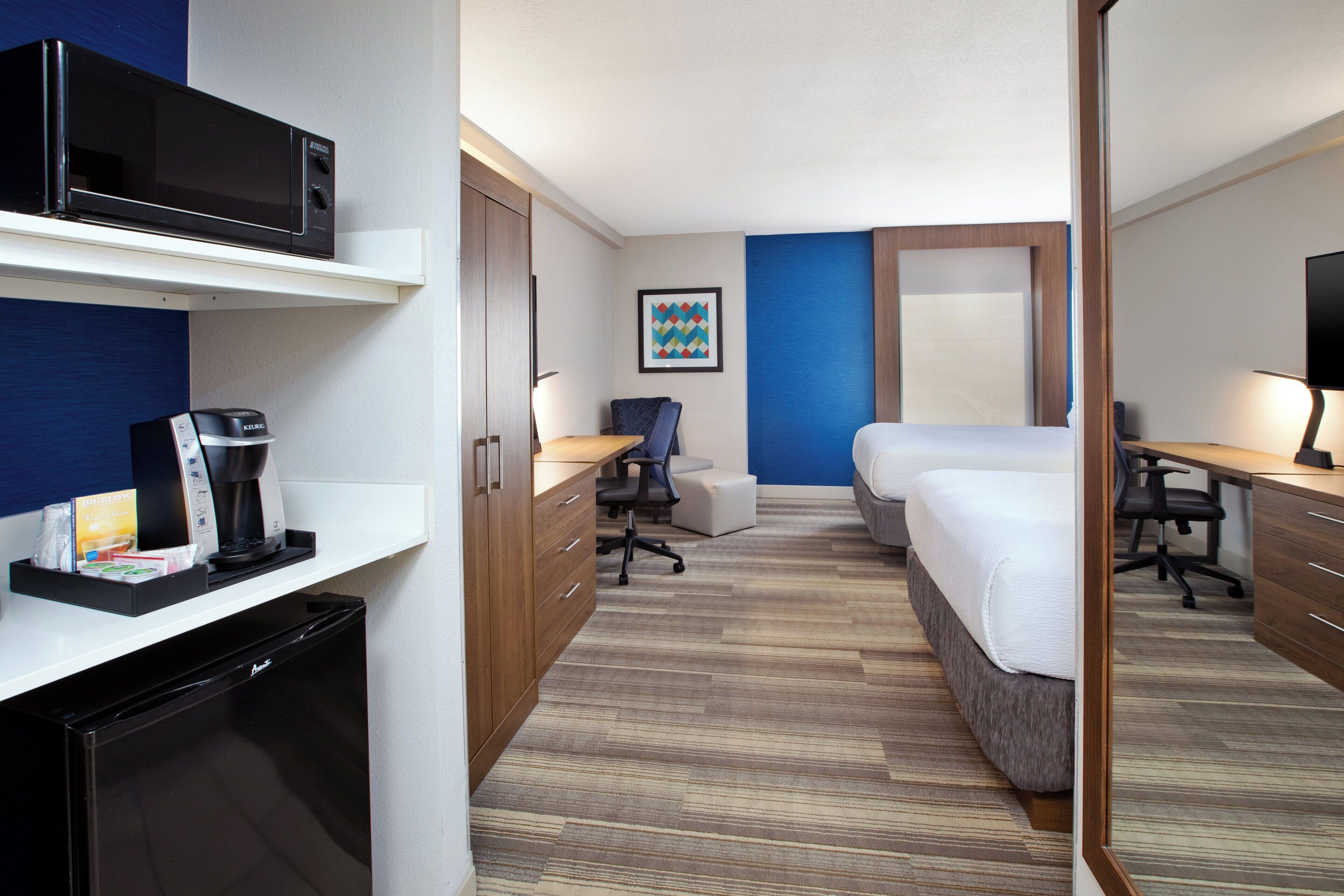 A hotel room with a microwave, coffee maker, desk, chair, and a bed.
