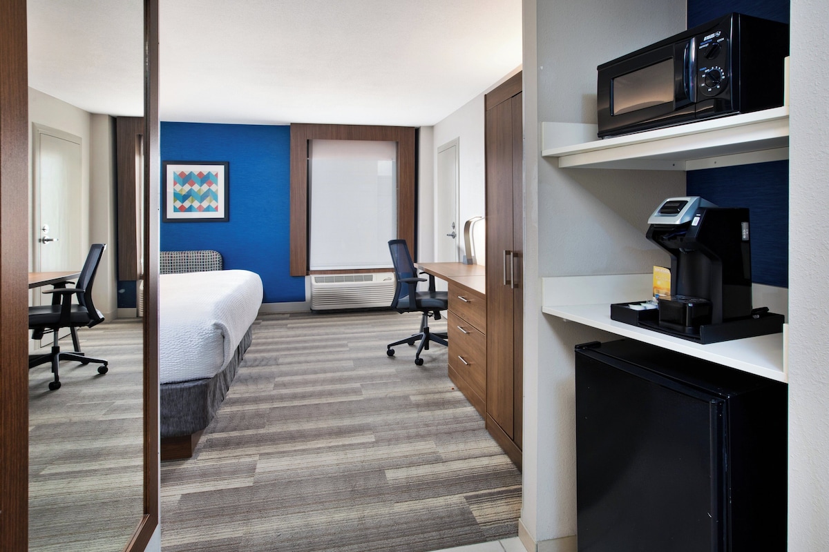 Habitación de hotel con cama, escritorio, sillas, microondas, cafetera y un refrigerador pequeño.