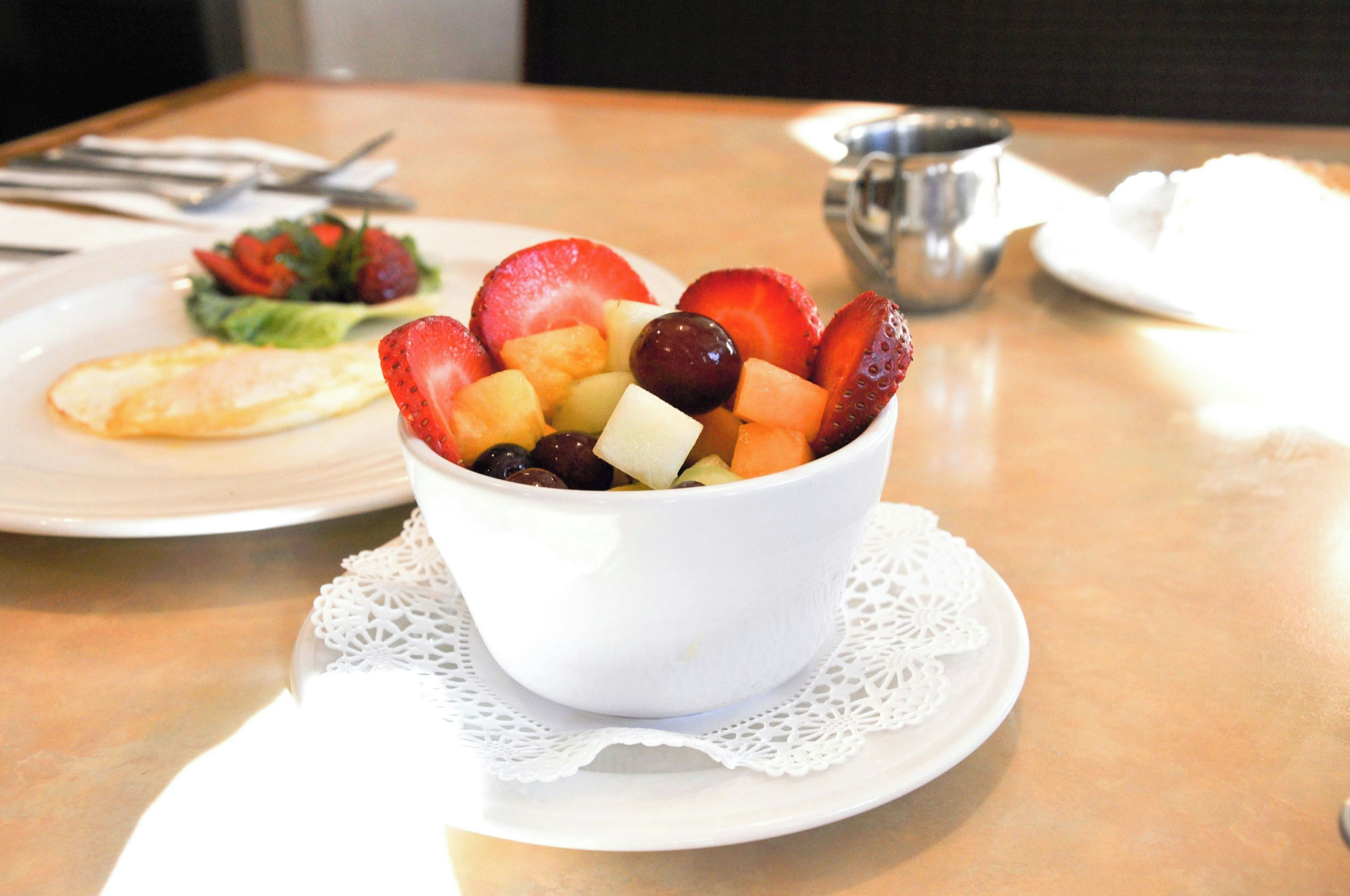Eine weiße Schüssel mit Obst auf einem Tisch mit Unterlage, ein Teller mit Salat und eine Tasse im Hintergrund.