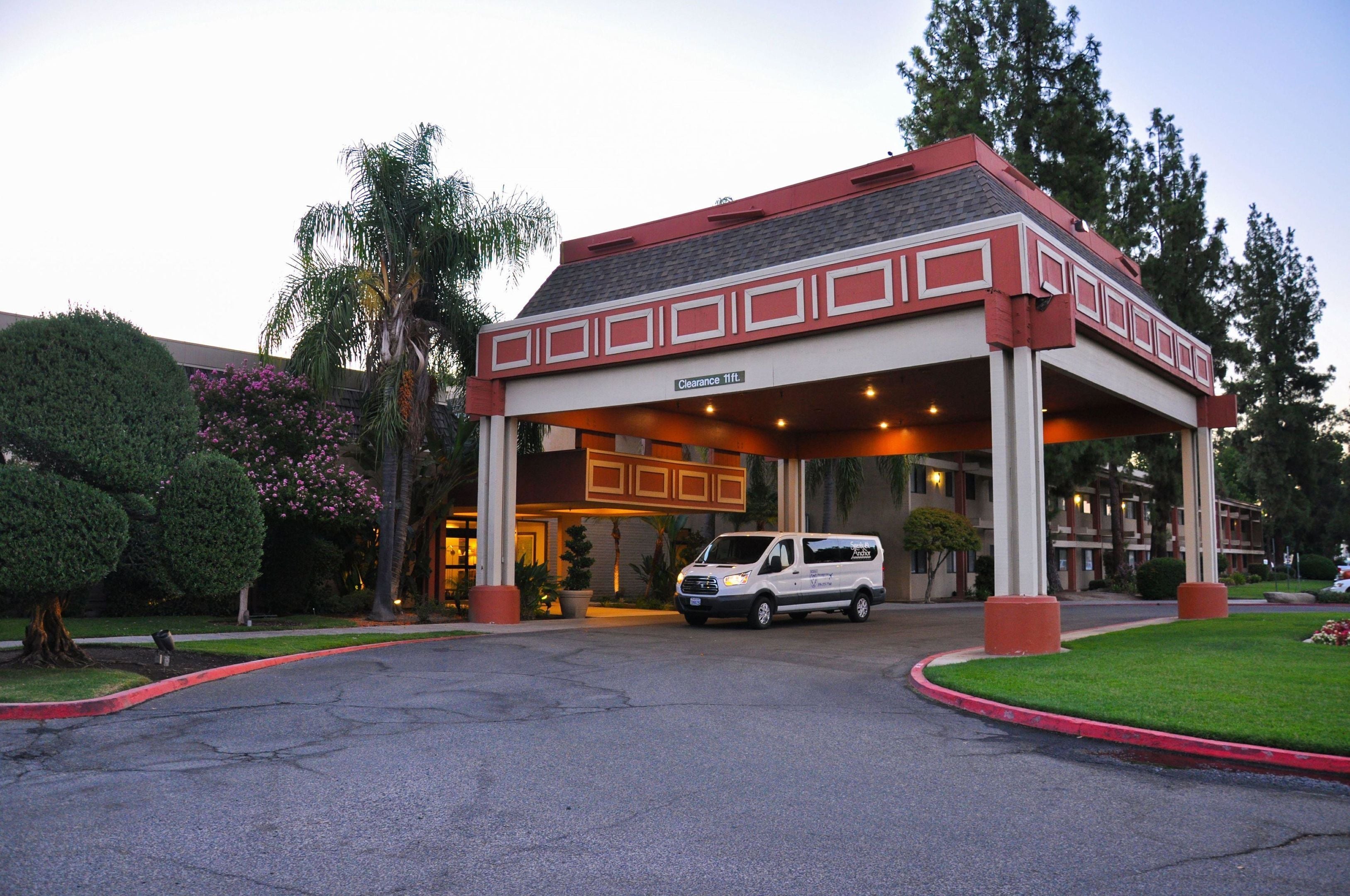 Entrada de hotel com área de espera coberta, uma van estacionada na frente e céu claro.