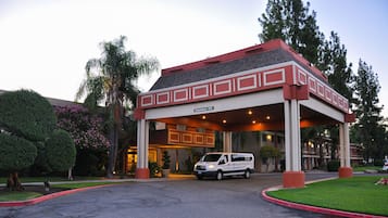 Entrada de hotel com área de espera coberta, uma van estacionada na frente e céu claro.