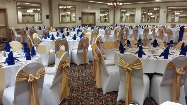Un salón de banquetes con mesas dispuestas para un evento formal, con manteles blancos y sillas adornadas con cintas doradas y azules.