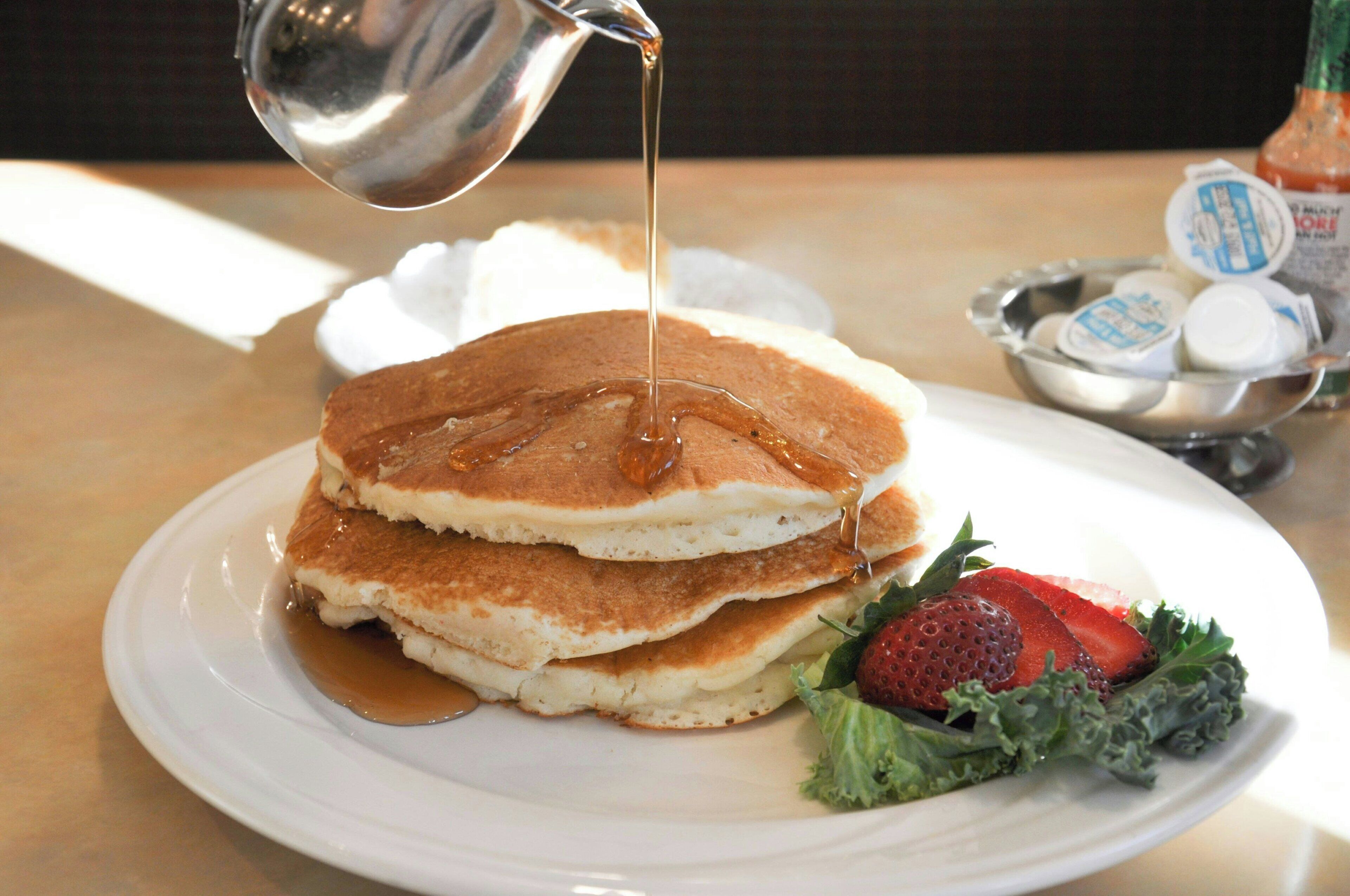 A stack of pancakes with syrup being poured on top, accompanied by a side of fresh strawberries and greens.