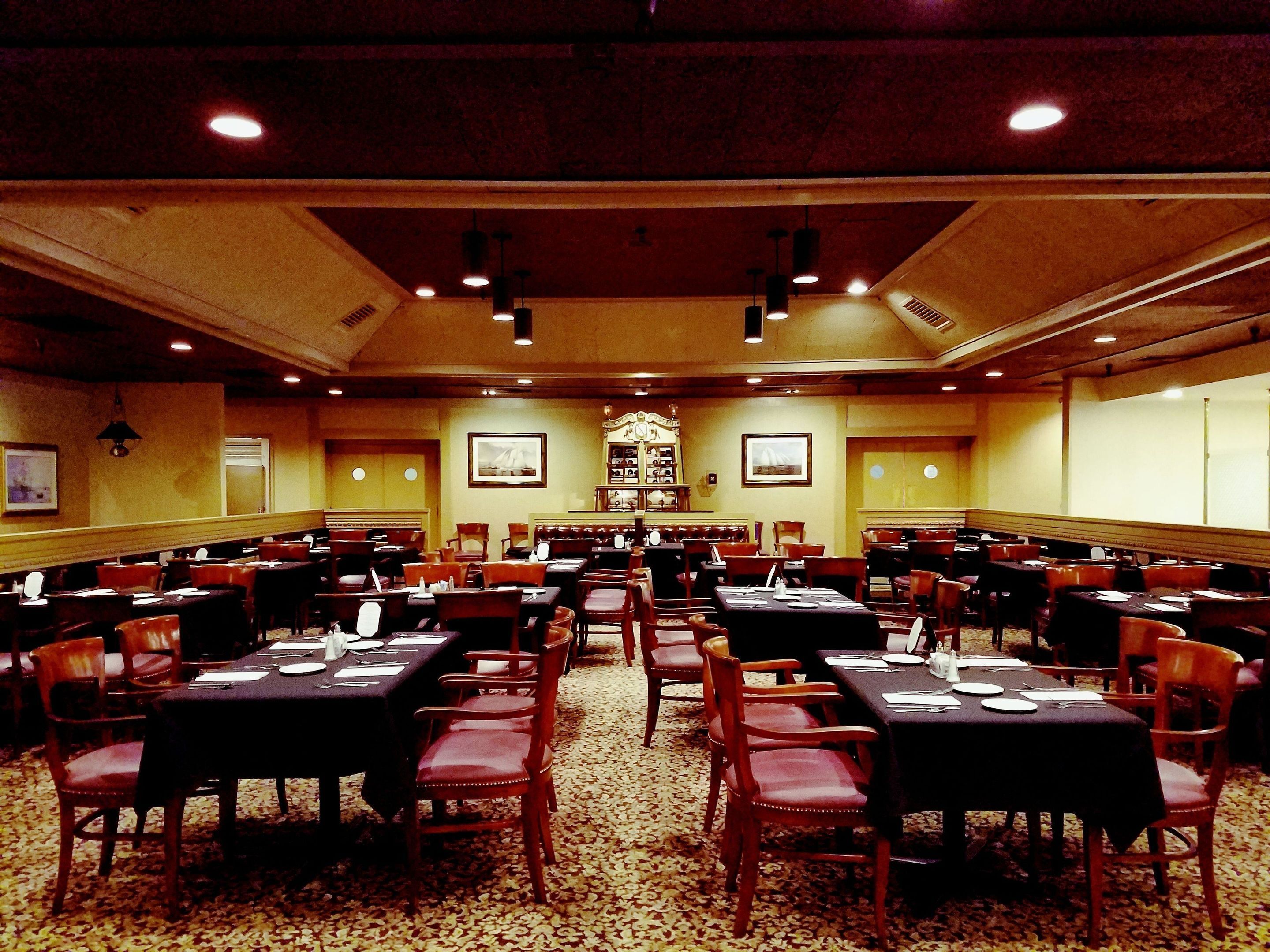 A large banquet hall with multiple tables and chairs, a high ceiling with recessed lighting, and framed pictures on the walls.