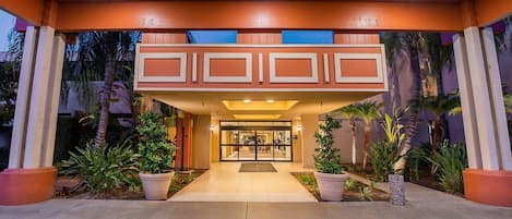 Una entrada de hotel con un corredor cubierto, plantas en macetas y puerta de vidrio.