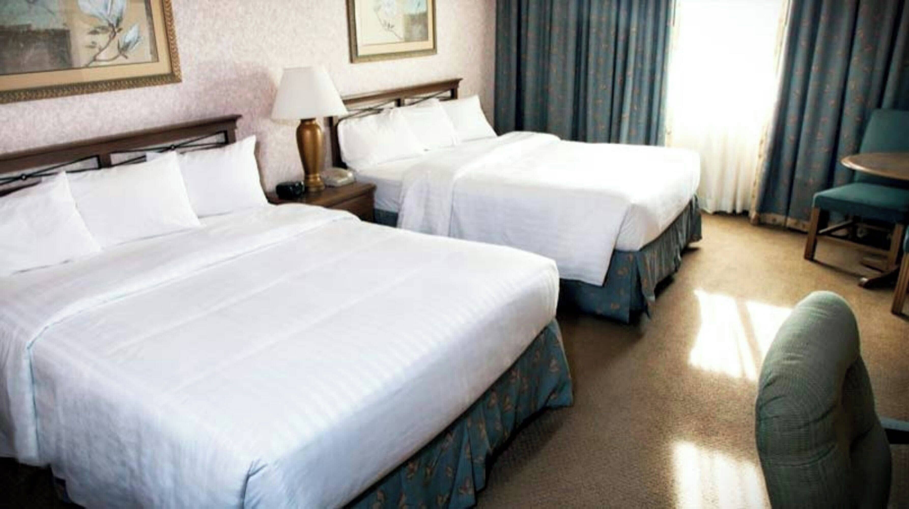 Une chambre d’hôtel avec deux lits, une table de chevet, une lampe, une chaise et une fenêtre avec des rideaux.