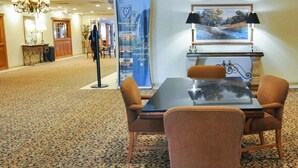Un hall de hotel con piso alfombrado, una mesa con sillas, un banderín azul, un cuadro enmarcado y un candelabro.