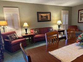 Uma sala de estar com sofá, cadeiras, mesa, luminárias e um quadro na parede.