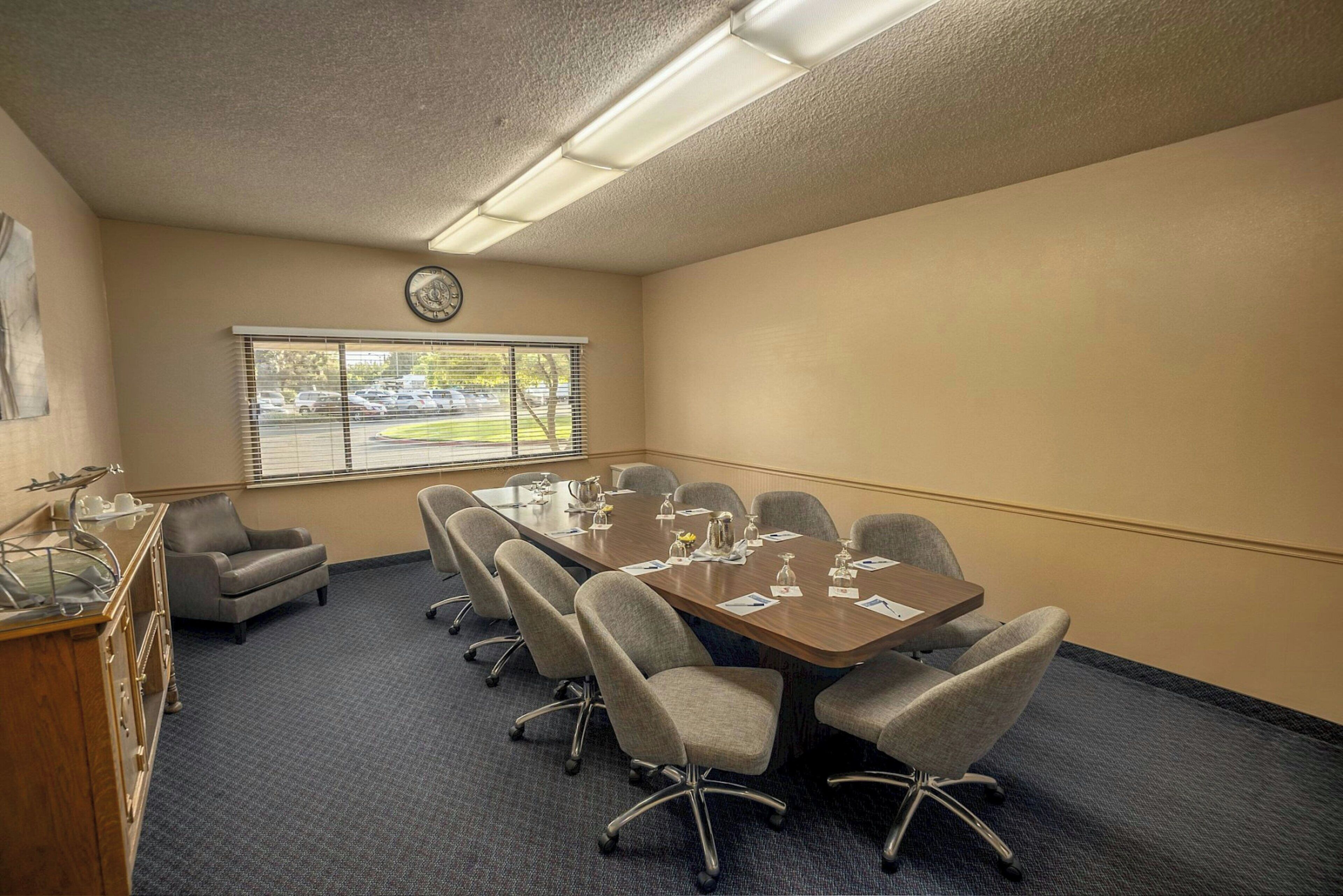 A conference room with a large table, six chairs, a clock on the wall, and a window with blinds.