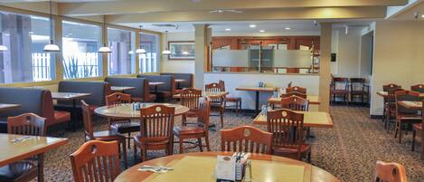 A hotel dining area with multiple tables and chairs, large windows, and ceiling fans.