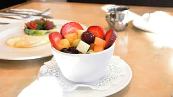 Un tazón blanco con fruta sobre una mesa, un mantel individual, un plato con ensalada y una taza al fondo.