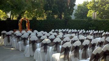 Rows of chairs with white covers and dark sashes, arranged outdoors with a backdrop of trees and a hedge.