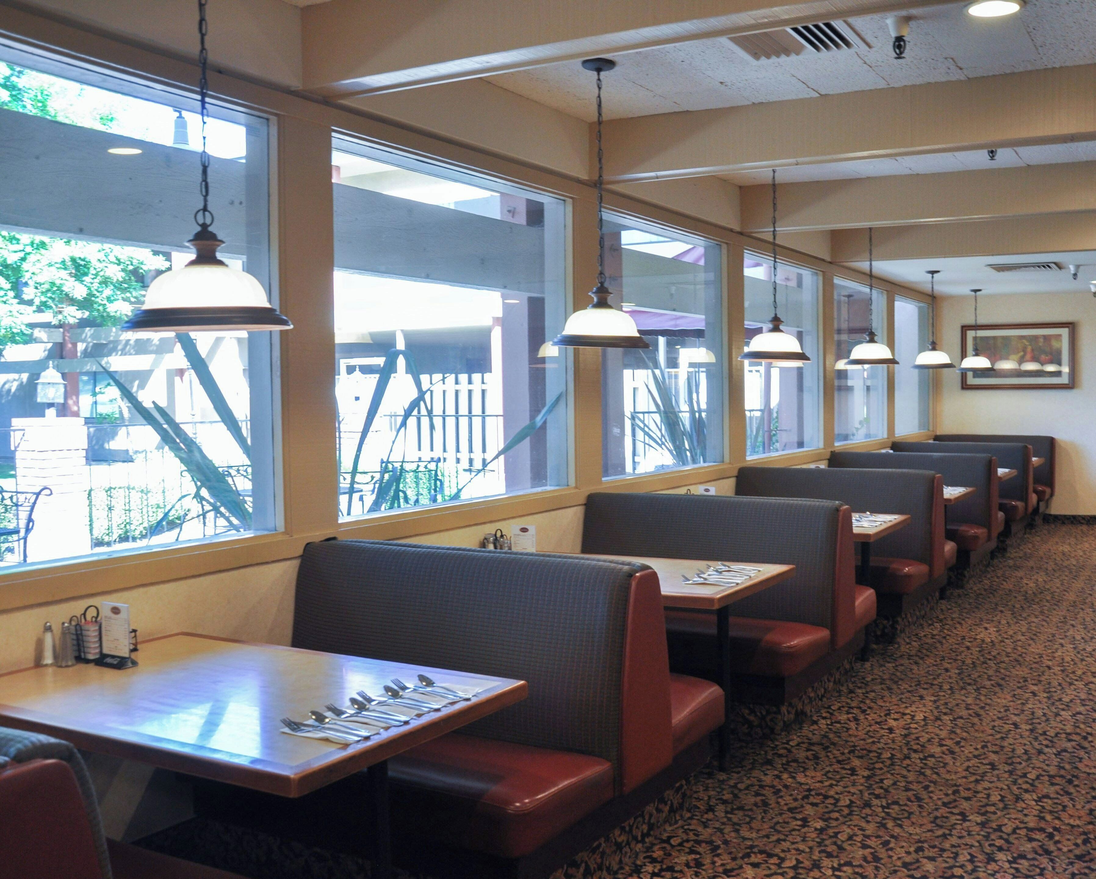 Una hilera de puestos con mesas, cada uno con cubiertos y servilletas, en un restaurante con ventanas grandes e iluminación colgante.