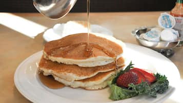 A stack of pancakes with syrup being poured on top, accompanied by a side of fresh strawberries and greens.
