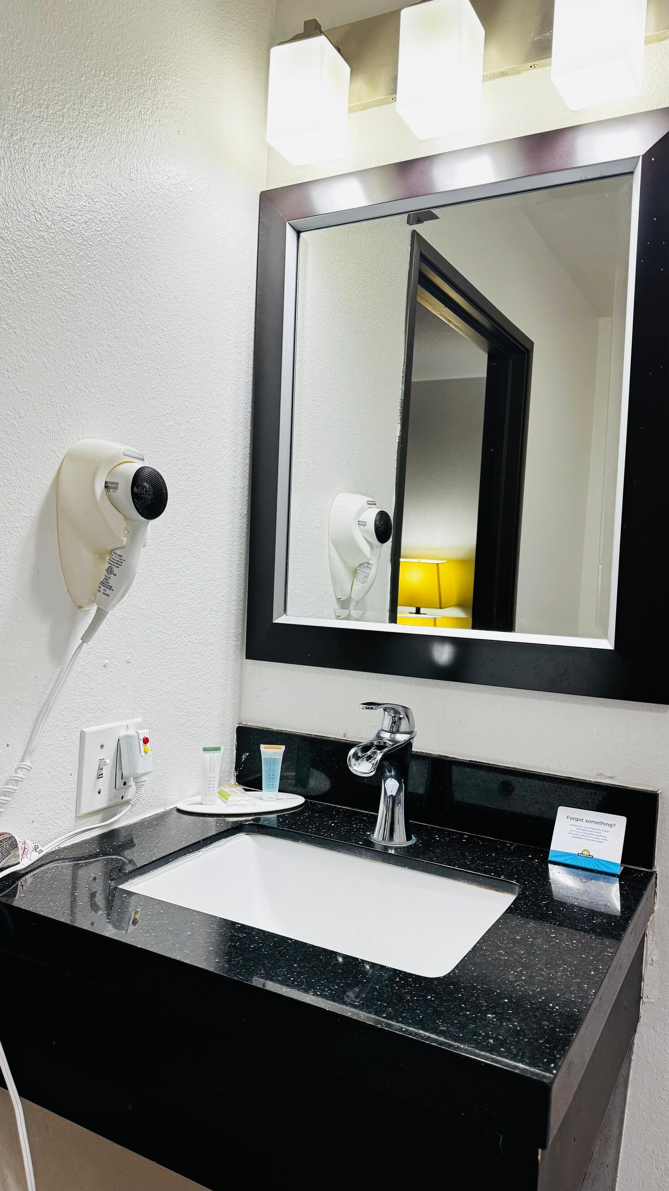Casa de banho de hotel com lavatório em bancada preta, espelho, secador de cabelo, telefone e dispensador de cartões.