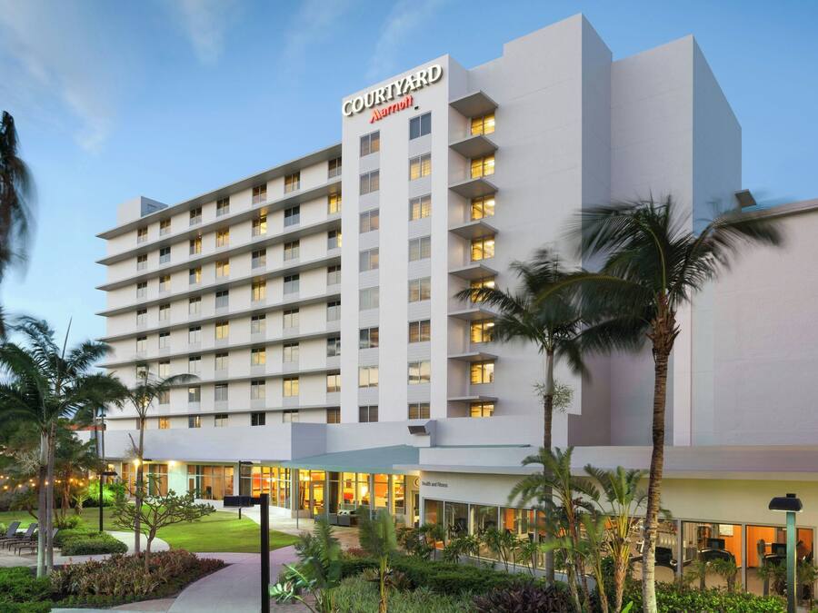 Un hotel Courtyard Marriott con varios pisos, un área exterior bien cuidada con palmeras y una entrada visible.