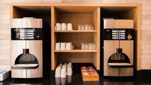 Una zona de cafè amb dues màquines d'espresso, una estant amb tasses, vasos i una caixa de llibres.