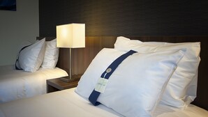 A hotel room with two beds, each with white linens and pillows, a bedside table with a lamp, and a dark wooden headboard.