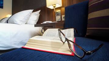 A hotel room with a bed, a bedside table with a lamp, and an open book with glasses on it.