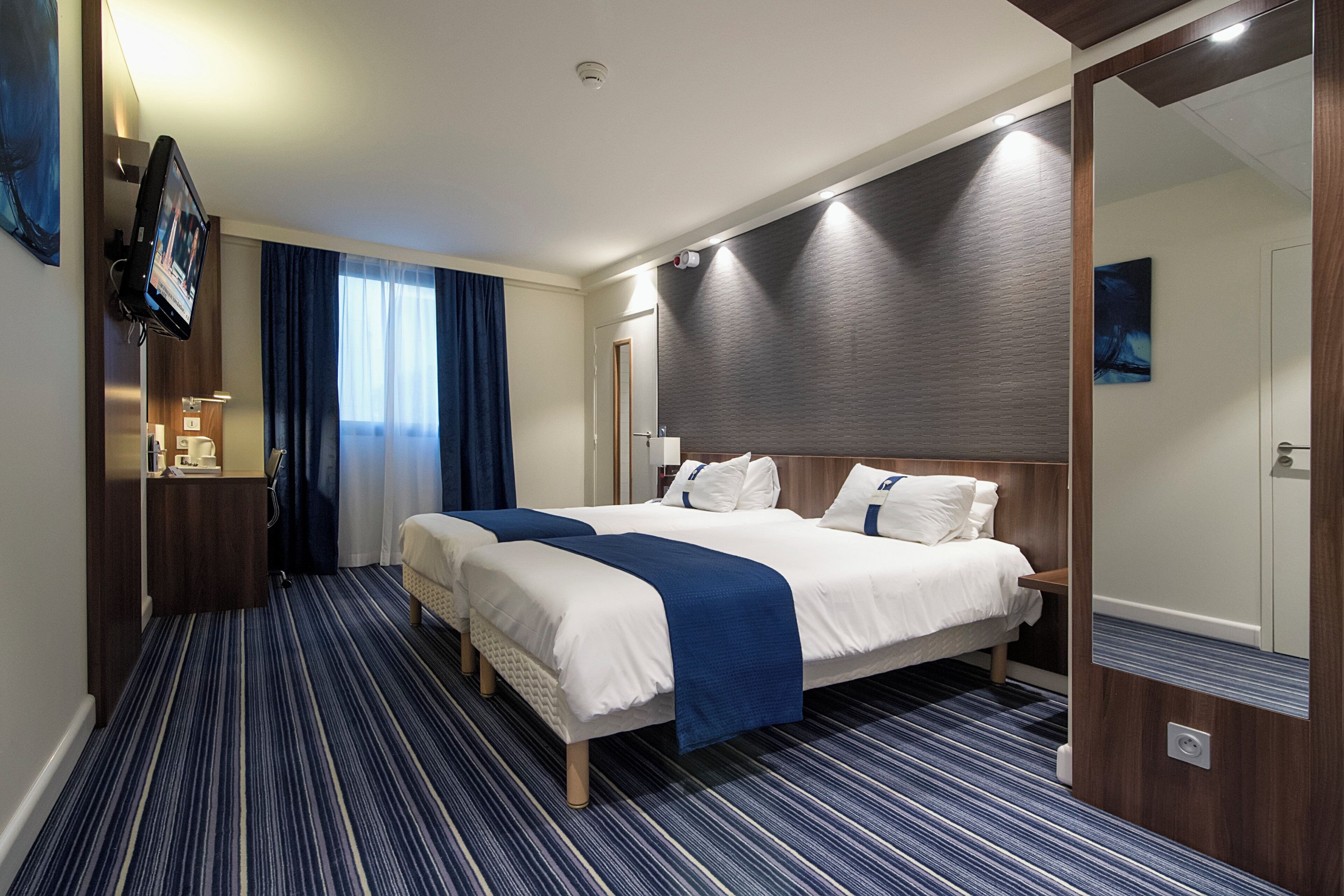 Habitación de hotel con una cama grande, televisión, espejo y ventana con cortinas.