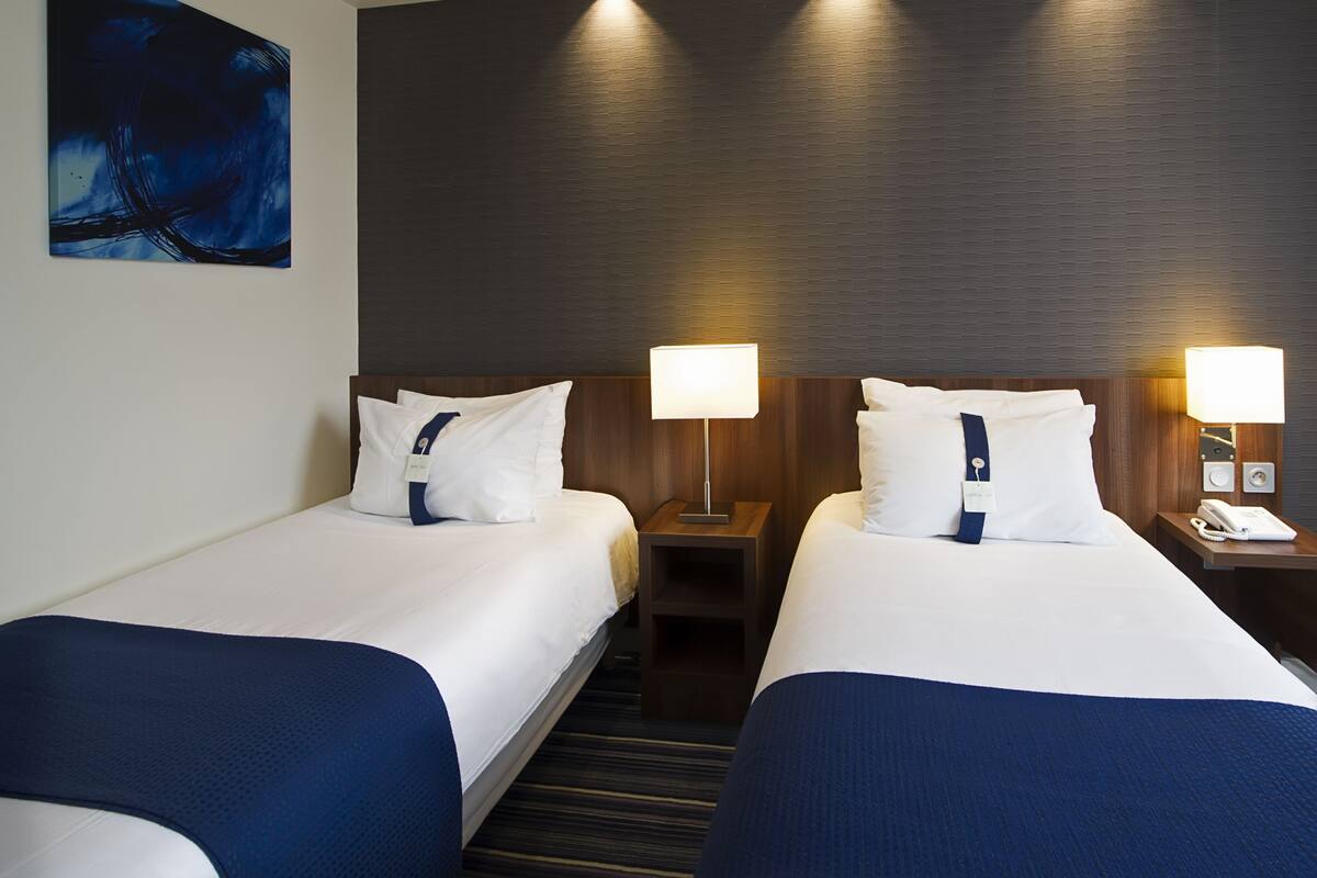 Habitación de hotel con dos camas, cada una con almohada y lámpara. Hay un cuadro en la pared y un cabecero de madera.