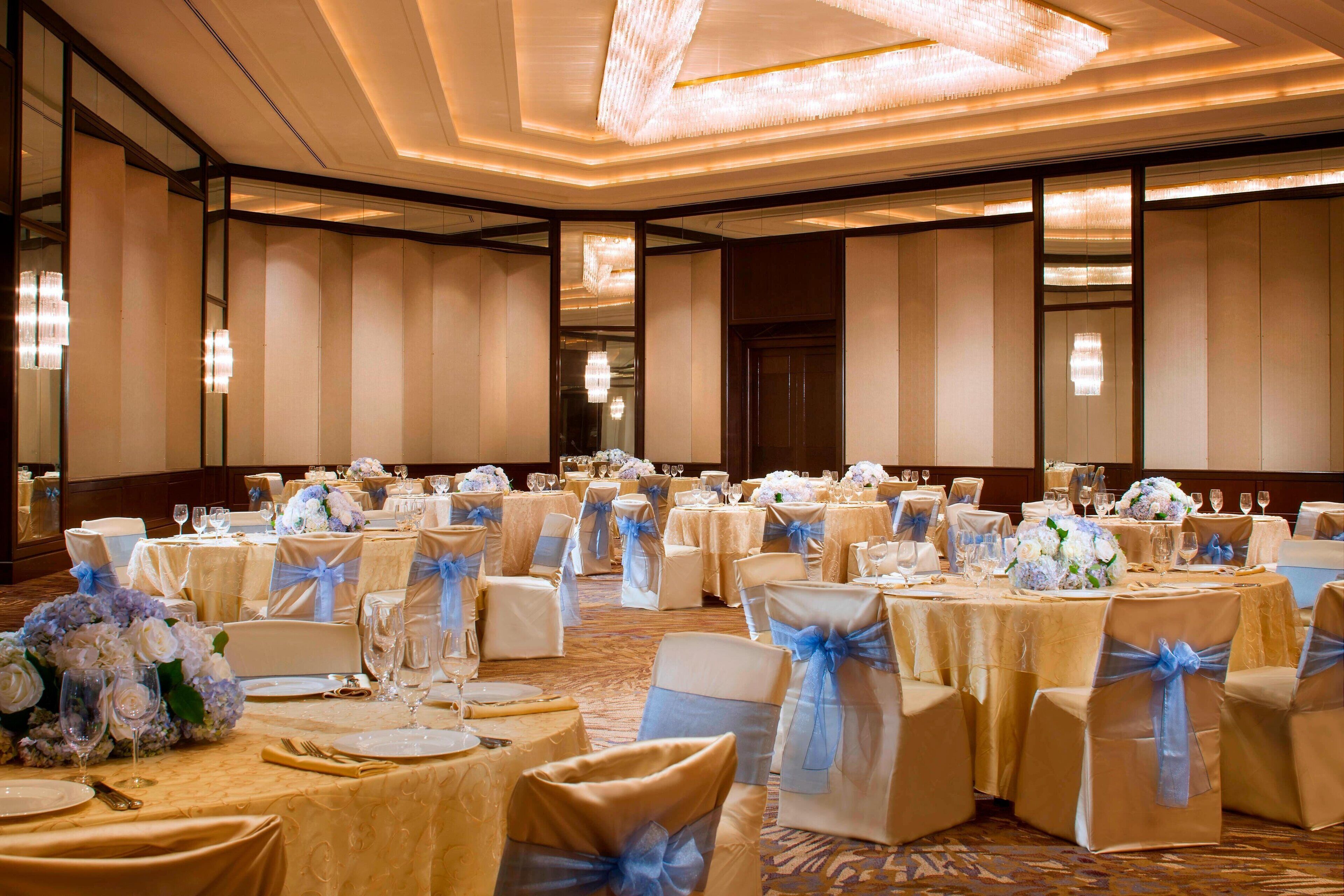A banquet hall with round tables set for a formal event, featuring white and gold tablecloths, chairs with blue sashes, and floral centerpieces.