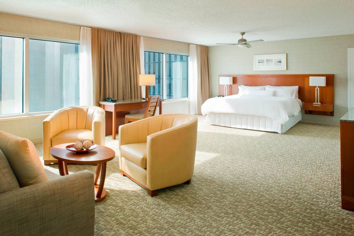 A hotel room with a bed, two armchairs, a small table with a tray, a desk with a lamp, and a ceiling fan.