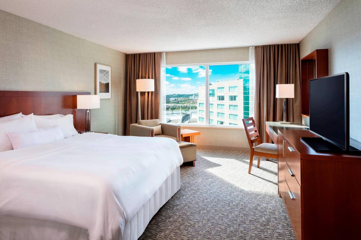 A hotel room with a large bed, a desk with a TV, a chair, and a view of the city through the window.