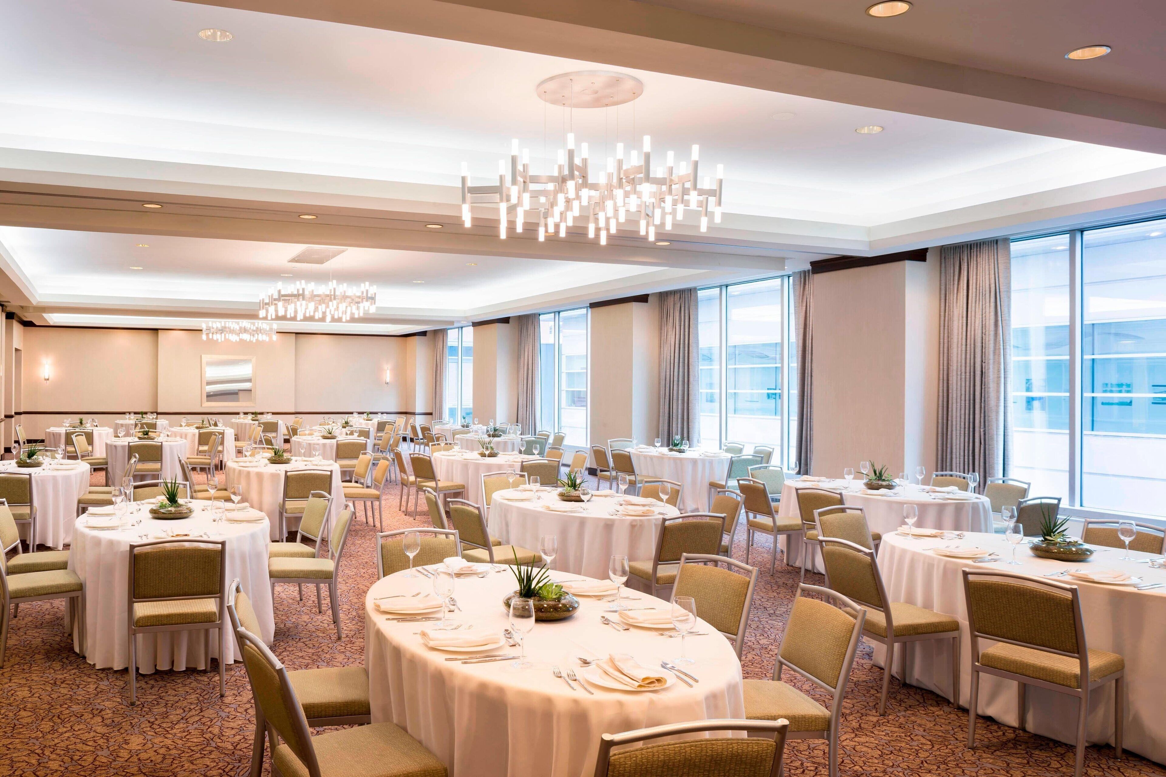 A banquet hall with round tables set for dining, large windows, and a modern chandelier.