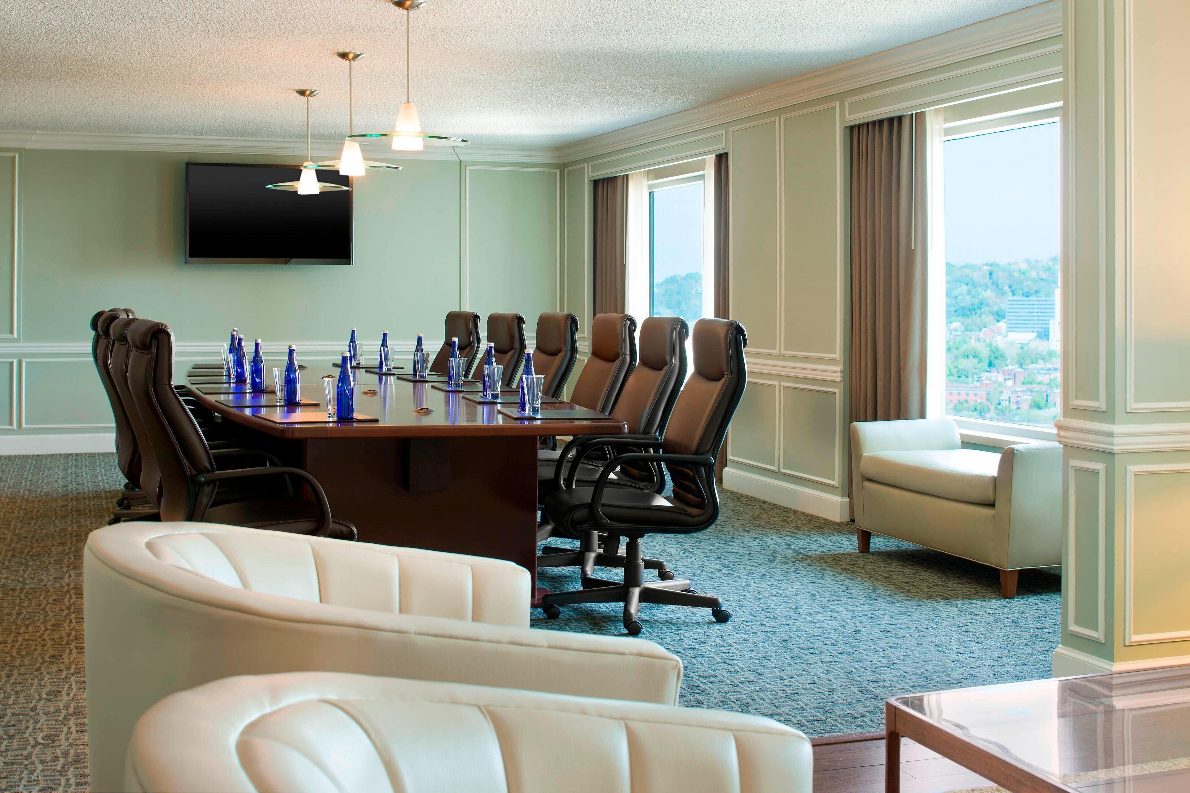 A conference room with a long table, multiple chairs, a TV on the wall, and a view through large windows.