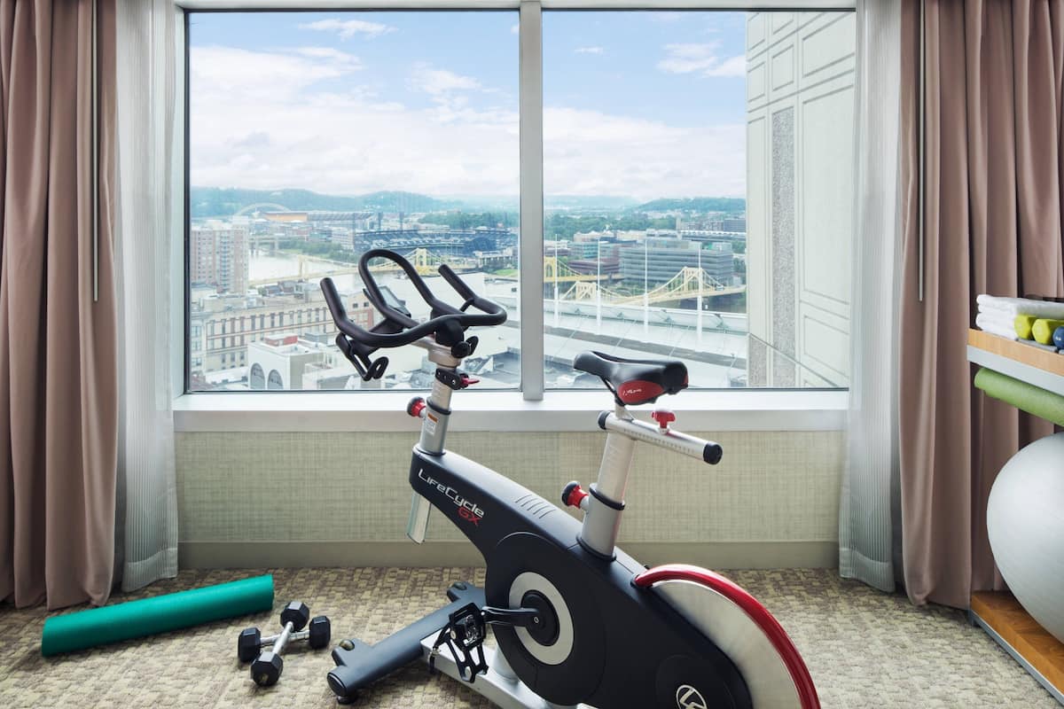 A room with a stationary bike, a pair of dumbbells, a yoga mat, and a view of a cityscape through a large window.