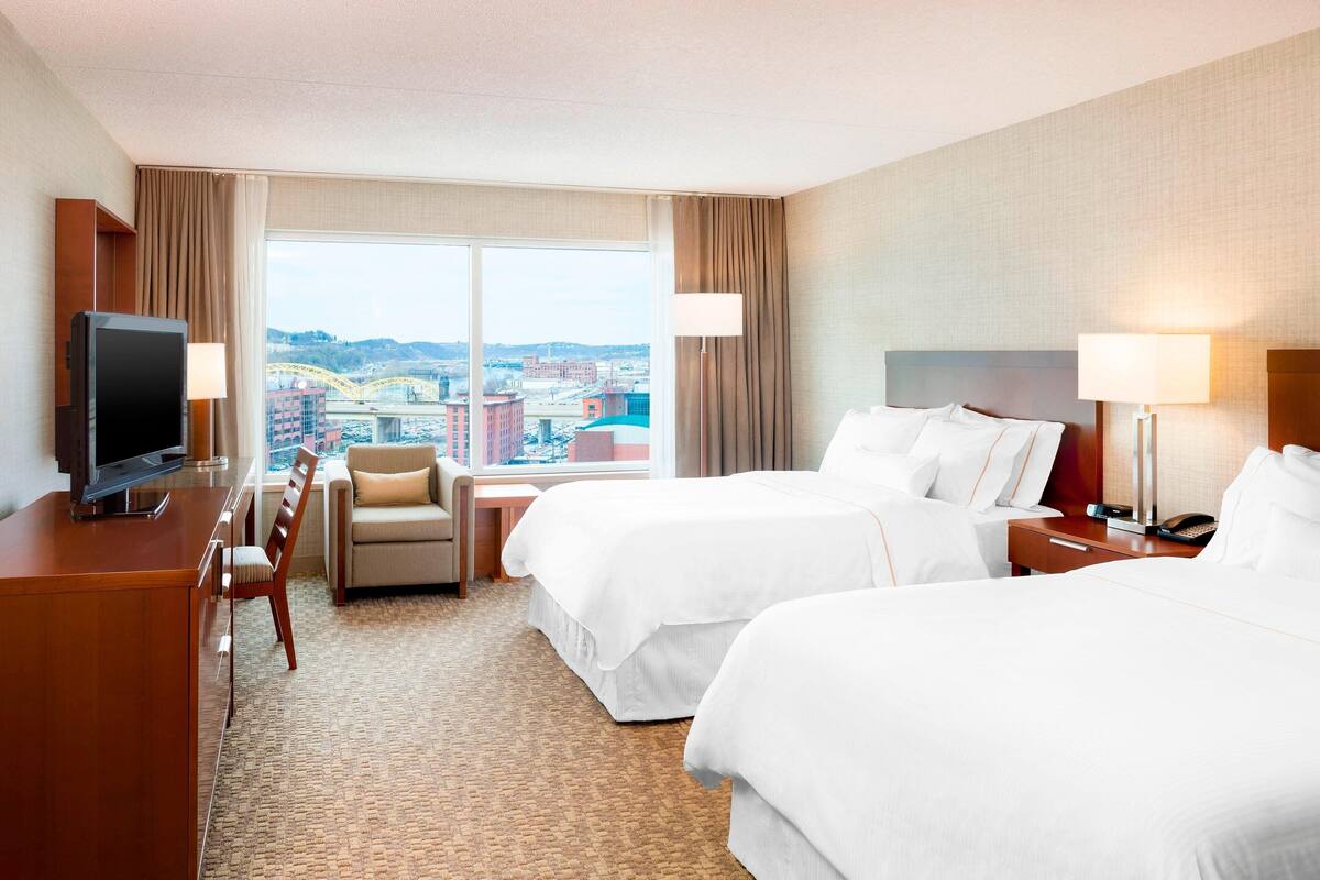 A hotel room with two beds, a desk, a TV, and a view of the city.