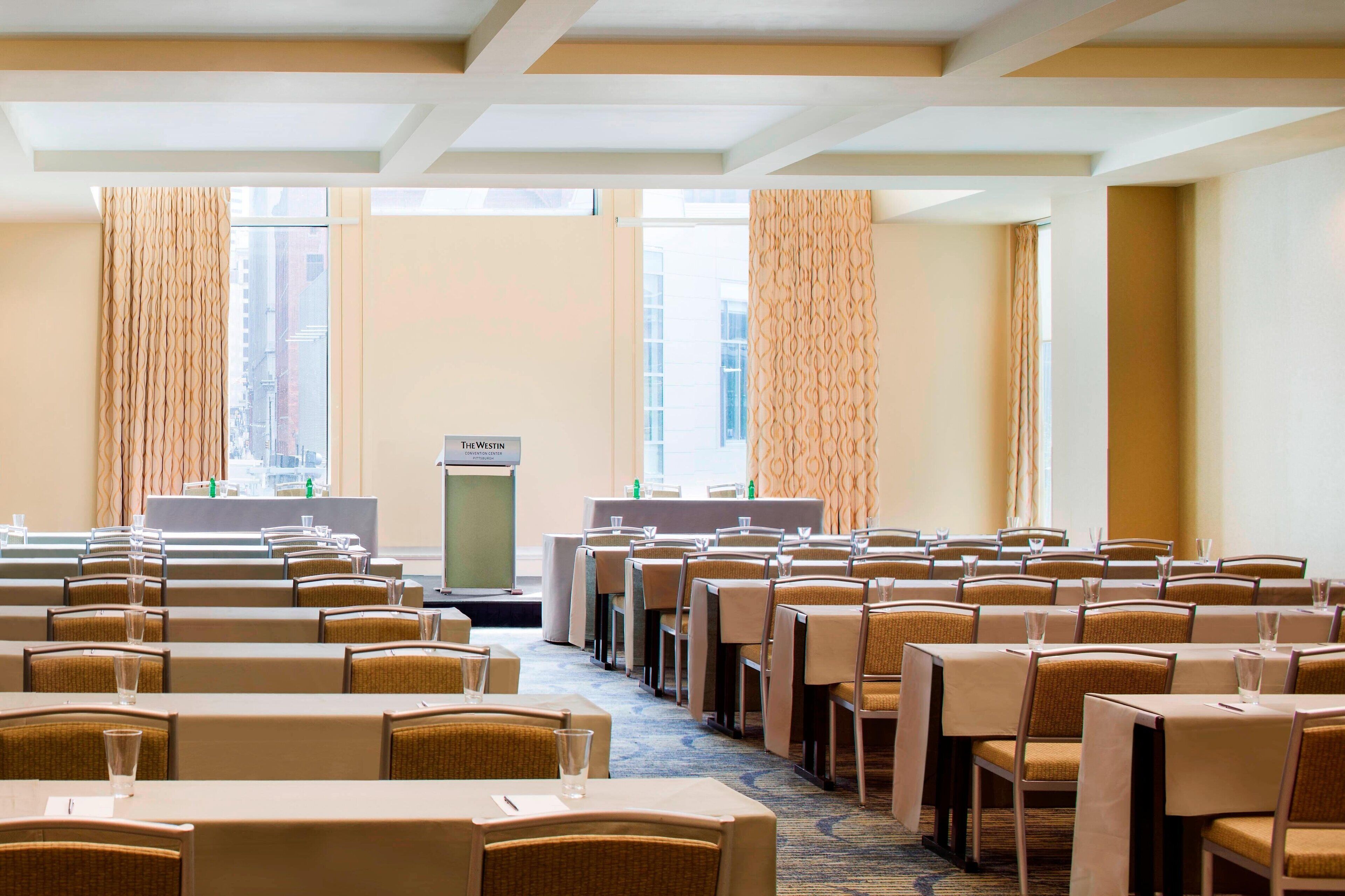 Un salón de conferencias con filas de mesas y sillas, un atril y ventanas grandes con cortinas.