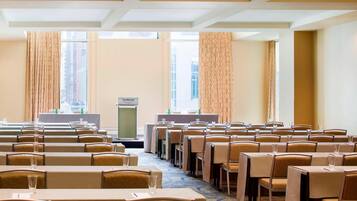Un salón de conferencias con filas de mesas y sillas, un atril y ventanas grandes con cortinas.